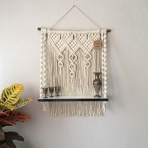 Puede incluir: Estante de pared de macramé color crema con un estante negro y objetos decorativos. El macramé presenta patrones de diamantes y cuentas de madera. Un pequeño jarrón y tres copas se exhiben en el estante.