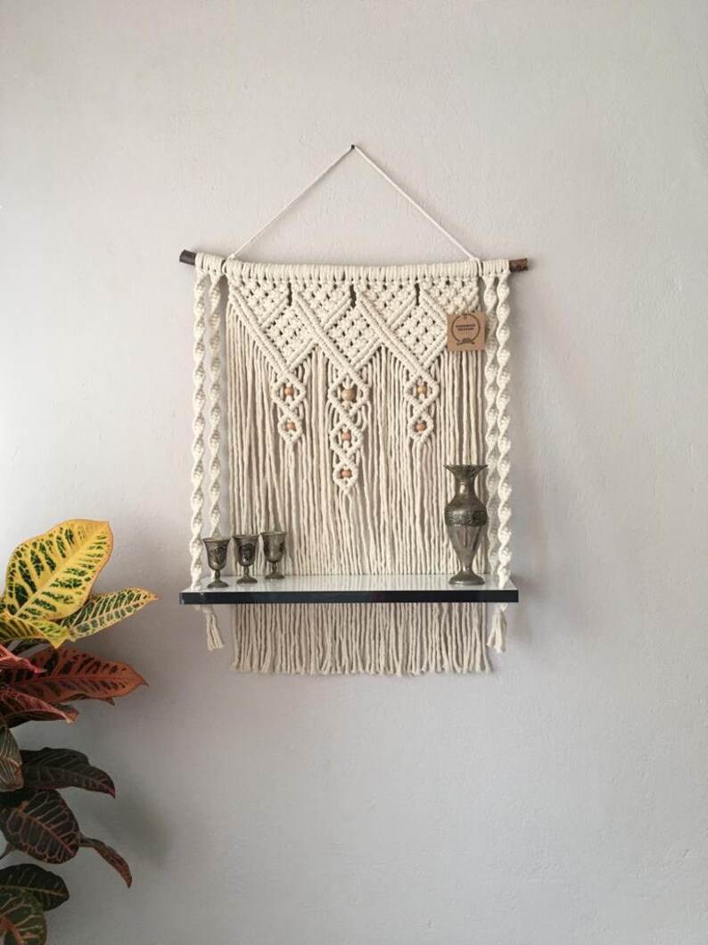 May include: Cream-colored macrame wall shelf with a black shelf and decorative items. The macrame features diamond patterns and wooden beads. A small vase and three goblets are displayed on the shelf.