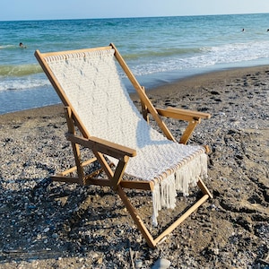 Puede incluir: Una silla de playa de madera con asiento y respaldo de macramé blanco. La silla está colocada en una playa de arena con el océano al fondo. La silla está plegada y lista para usar.