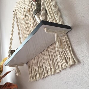 May include: A macrame hanging shelf with a black shelf and cream-colored macrame detailing. The shelf is attached to the wall and has a decorative item on top. The macrame design includes knotted patterns and fringe.