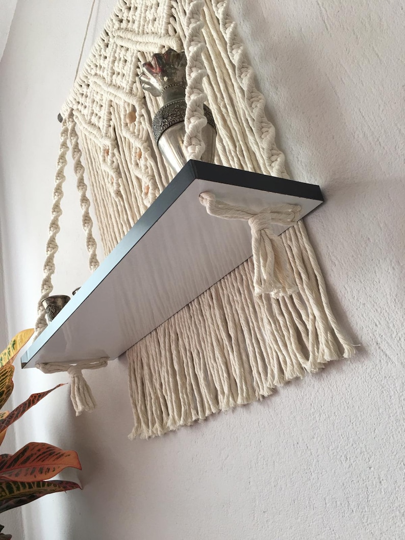 May include: A macrame hanging shelf with a black shelf and cream-colored macrame detailing. The shelf is attached to the wall and has a decorative item on top. The macrame design includes knotted patterns and fringe.