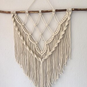 May include: A white macrame wall hanging with a diamond pattern and fringe. The hanging is attached to a natural wood branch.