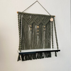 May include: Olive green macrame wall shelf with a white shelf and black trim. The macrame features diamond patterns and wooden beads. A wooden dowel and hanging cord support the shelf.
