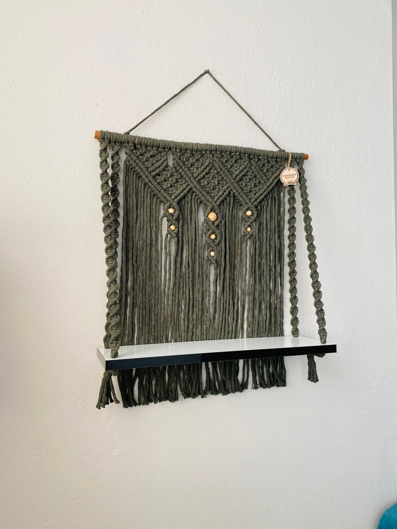 May include: Olive green macrame wall shelf with a white shelf and black trim. The macrame features diamond patterns and wooden beads. A wooden dowel and hanging cord support the shelf.