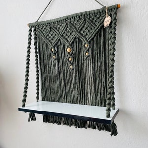 May include: A macrame wall shelf in a muted green color. The shelf features a woven design with wooden beads and a white shelf. The shelf is suspended by a rope and is suitable for displaying small items.