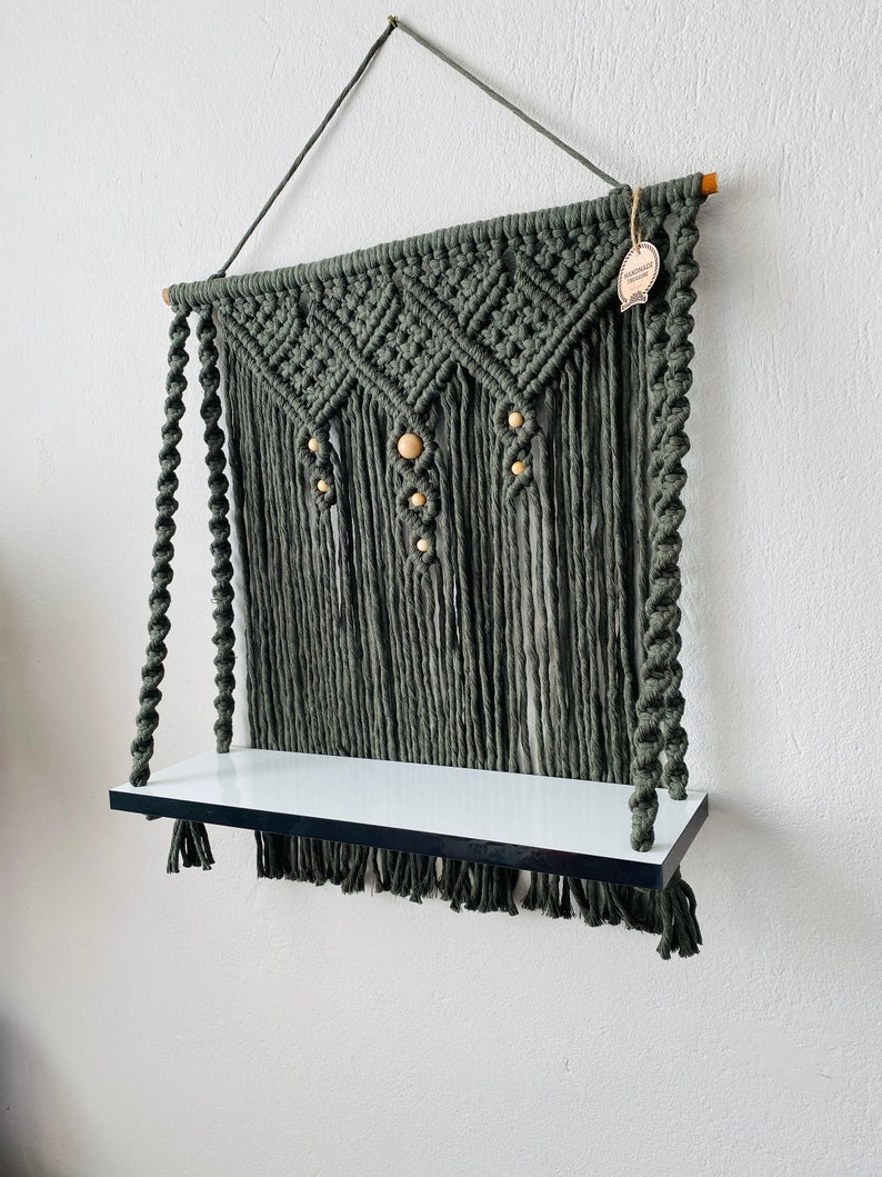 May include: A macrame wall shelf in a muted green color. The shelf features a woven design with wooden beads and a white shelf. The shelf is suspended by a rope and is suitable for displaying small items.