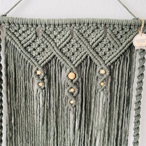May include: Sage green macrame wall hanging with a geometric design and wooden beads. The handmade piece features intricate knotting and fringe, suspended from a wooden dowel. A tag that says "HANDMADE TREASURE" is attached.