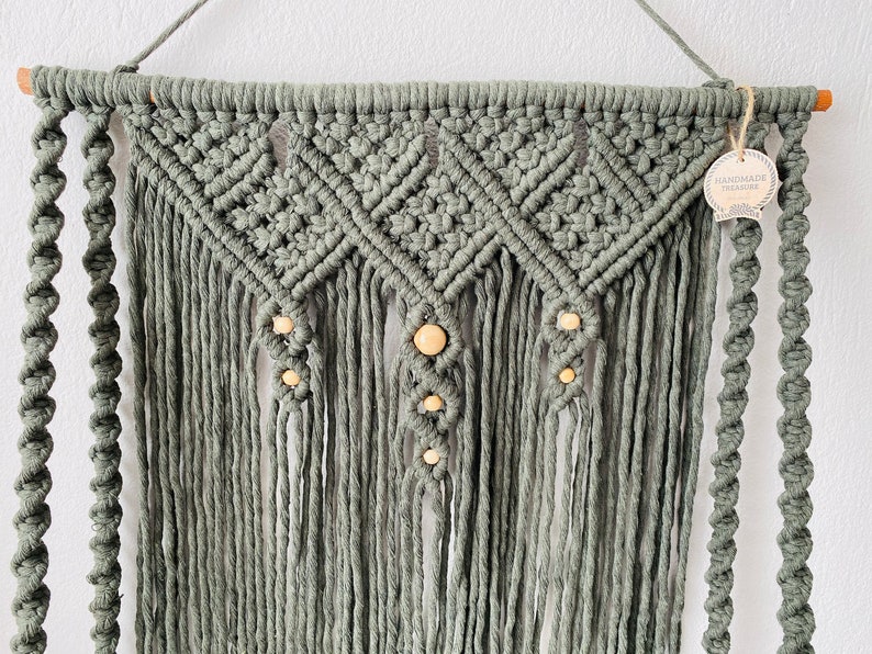 May include: Sage green macrame wall hanging with a geometric design and wooden beads. The handmade piece features intricate knotting and fringe, suspended from a wooden dowel. A tag that says "HANDMADE TREASURE" is attached.