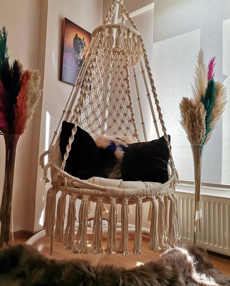 Puede incluir: Una silla colgante de macram&eacute; color crema con cojines negros y un coj&iacute;n decorativo de piel. La silla est&aacute; suspendida del techo y tiene una base con flecos. Altos jarrones con plantas secas de colores a ambos lados.