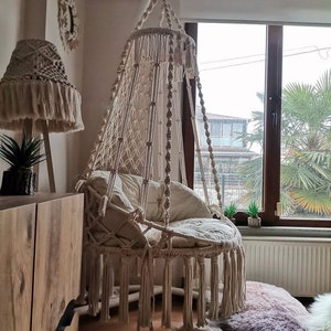 Puede incluir: Una silla colgante de macram&eacute; color crema con asiento redondo y flecos. La silla est&aacute; suspendida del techo cerca de una ventana. Tambi&eacute;n se ven una pantalla de l&aacute;mpara de macram&eacute; a juego y cojines decorativos.