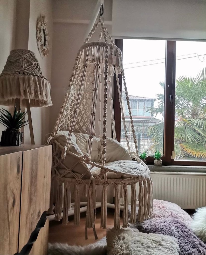 Puede incluir: Una silla colgante de macram&eacute; color crema con asiento redondo y flecos. La silla est&aacute; suspendida del techo cerca de una ventana. Tambi&eacute;n se ven una pantalla de l&aacute;mpara de macram&eacute; a juego y cojines decorativos.