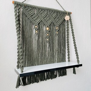 May include: A handmade macrame wall shelf in a muted green color. The shelf features a woven design with wooden beads and a white shelf with a black border. The macrame has a fringe at the bottom and is suspended by a rope.