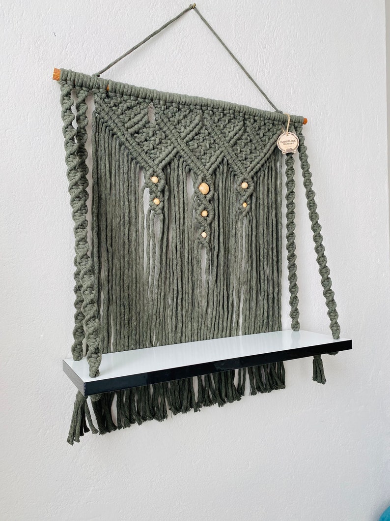 May include: A handmade macrame wall shelf in a muted green color. The shelf features a woven design with wooden beads and a white shelf with a black border. The macrame has a fringe at the bottom and is suspended by a rope.