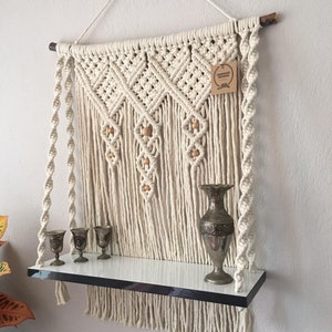 May include: Cream-colored macrame wall shelf with a white shelf and a silver metal trim. The shelf holds three small silver goblets and a decorative silver vase. The macrame design includes wooden beads and a hanging tag.
