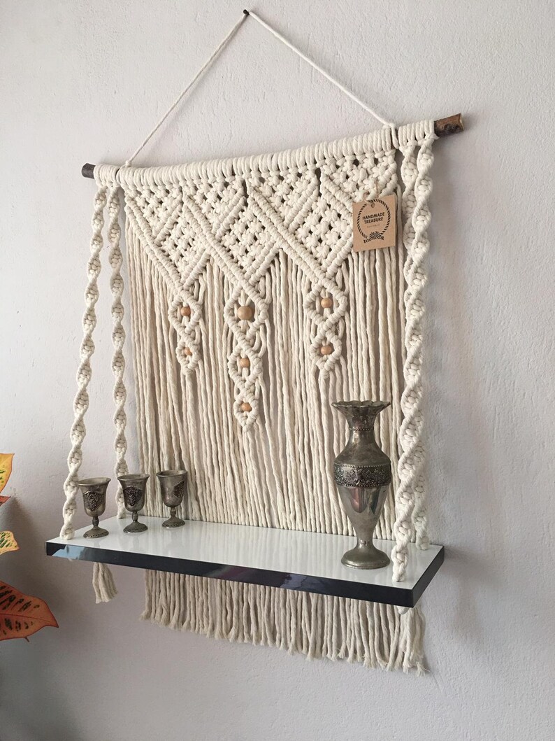 May include: Cream-colored macrame wall shelf with a white shelf and a silver metal trim. The shelf holds three small silver goblets and a decorative silver vase. The macrame design includes wooden beads and a hanging tag.