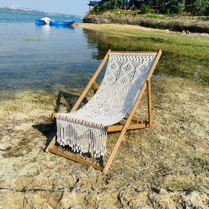 Puede incluir: Una silla de playa de macramé con marco de madera y asiento tejido blanco. La silla está colocada en una playa de arena cerca del agua. Dos botes azules se ven al fondo.