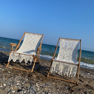 Puede incluir: Dos sillas de playa de madera con asientos y flecos de macramé blanco en una playa arenosa. Las sillas tienen un marco de madera natural y están orientadas hacia el océano bajo un cielo azul. La playa está cubierta de arena y guijarros.