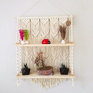 May include: A white macrame wall shelf with two wooden shelves. The shelves are decorated with small plants, a ladybug figurine, and dried flowers. The macrame is made with a knot pattern and has a fringe at the bottom.