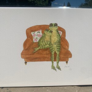May include: A watercolour painting of two green frogs sitting on a brown sofa with a floral patterned cushion. The frogs are looking up and the sofa has a brown wooden frame.