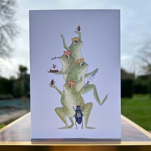 May include: A stack of four green frogs wearing paper crowns, each holding a different festive item. The bottom frog is holding a beetle with a gift box. The frog above is holding a test tube with purple glitter. The next frog is holding a plate with a Christmas pudding. The top frog is holding a red ladybug.