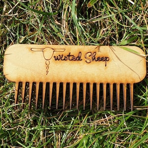 May include: A wooden comb with a stylised sheep design and the text "Twisted Sheep" engraved on the top. The comb is light brown and is lying on a bed of green grass.
