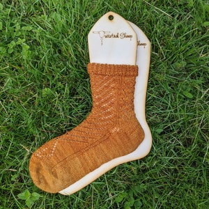 May include: A pair of brown knitted socks with a lacy pattern, displayed on a wooden sock blocker with the text "Twisted Sheep" on it.