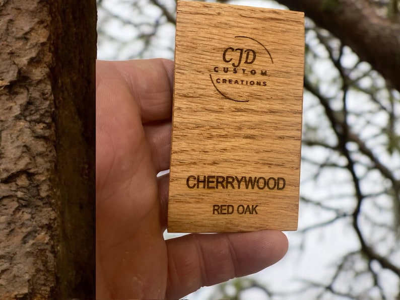 A stained wood sample swatch labeled "CHERRYWOOD" on "RED OAK" with a laser-engraved "CJD Custom Creations" logo, showing the stain color and grain pattern for reference