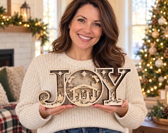 May include: A layered wooden cutout sign spelling the word JOY, featuring a detailed nativity scene inside the letter O. The design includes silhouettes of figures, animals, buildings, and stars, crafted from light and dark wood tones.