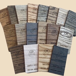 A collection of stained wood sample swatches arranged in a fan layout, each labeled with the stain color and wood type. Swatches include shades like Sedona, Cherry, Dark Walnut, Ebony, Slate, Red Mahogany, and Golden Oak, all engraved with the "CJD Custom Creations" logo. These samples show how different Minwax stains appear on Red Oak wood, helping customers choose the right finish for their woodworking or home improvement projects.
