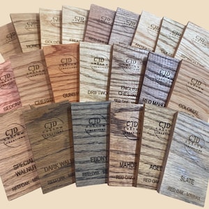 A collection of stained wood sample swatches arranged in a fan layout, each labeled with the stain color and wood type. Swatches include shades like Sedona, Cherry, Dark Walnut, Ebony, Slate, Red Mahogany, and Golden Oak, all engraved with the "CJD Custom Creations" logo. These samples show how different Minwax stains appear on Red Oak wood, helping customers choose the right finish for their woodworking or home improvement projects.