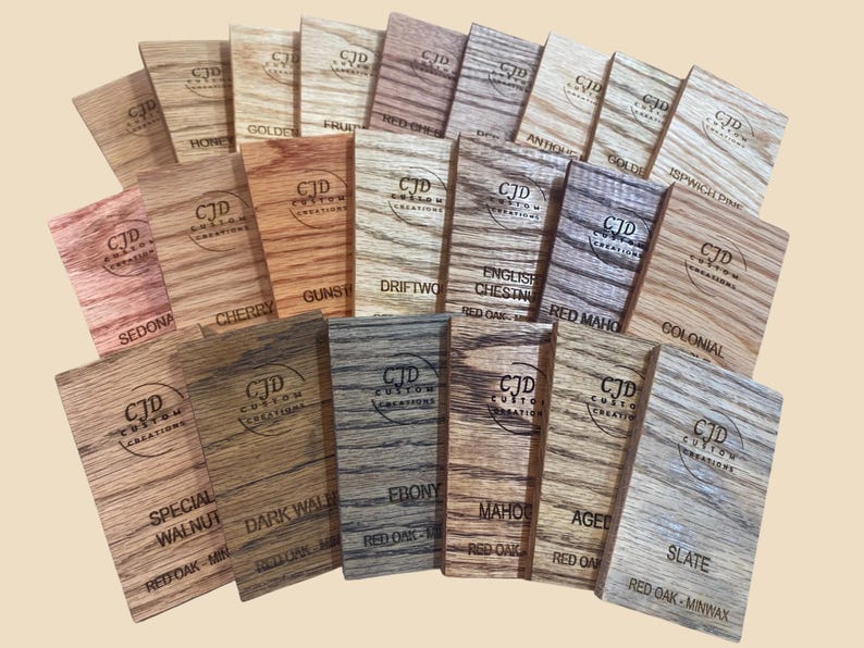 A collection of stained wood sample swatches arranged in a fan layout, each labeled with the stain color and wood type. Swatches include shades like Sedona, Cherry, Dark Walnut, Ebony, Slate, Red Mahogany, and Golden Oak, all engraved with the "CJD Custom Creations" logo. These samples show how different Minwax stains appear on Red Oak wood, helping customers choose the right finish for their woodworking or home improvement projects.
