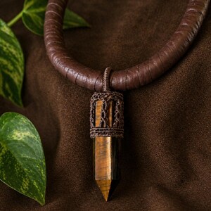 May include: A brown leather choker necklace with a pendant. The pendant is a tiger's eye crystal, encased in a brown woven design. The necklace is displayed on a brown textured surface, with green leaves in the background.
