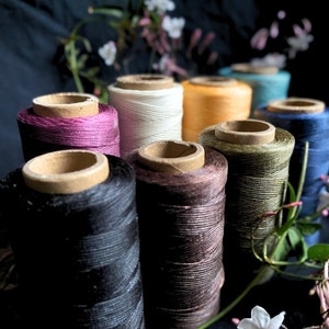 Könnte beinhalten: Eine Sammlung von acht Garnrollen in verschiedenen Farben, darunter Weiß, Braun, Schwarz, Rosa und Grün. Die Rollen sind in einem Halbkreis auf einem dunklen Hintergrund angeordnet.