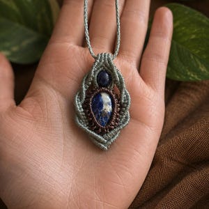 May include: A handmade macrame pendant necklace featuring a central oval blue stone with white inclusions, surrounded by a woven design in shades of blue and brown. The necklace has a light blue cord.