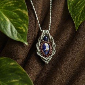 May include: A handmade macrame necklace featuring a blue and white gemstone pendant. The pendant is framed by a woven design in shades of green and brown, suspended from a matching cord. The necklace is displayed against a brown background.