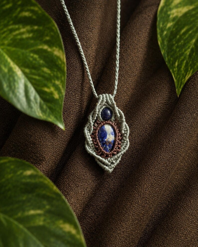 May include: A handmade macrame necklace featuring a blue and white gemstone pendant. The pendant is framed by a woven design in shades of green and brown, suspended from a matching cord. The necklace is displayed against a brown background.