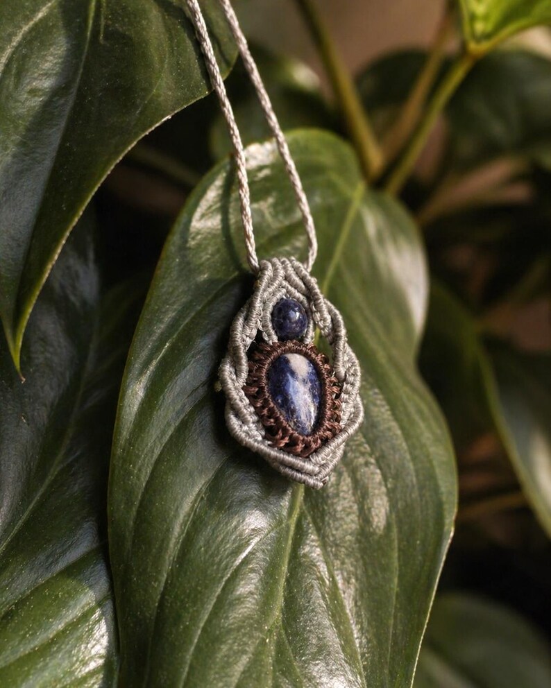 May include: A handmade macrame necklace featuring a blue gemstone pendant. The pendant is encased in a woven, gray macrame design with a brown border. The necklace hangs on a silver cord, set against a backdrop of green leaves.