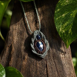 May include: A handmade macrame necklace featuring a blue and white stone pendant. The pendant is encased in a gray macrame design with a dark brown border. The necklace is displayed on a piece of weathered wood, surrounded by green foliage.
