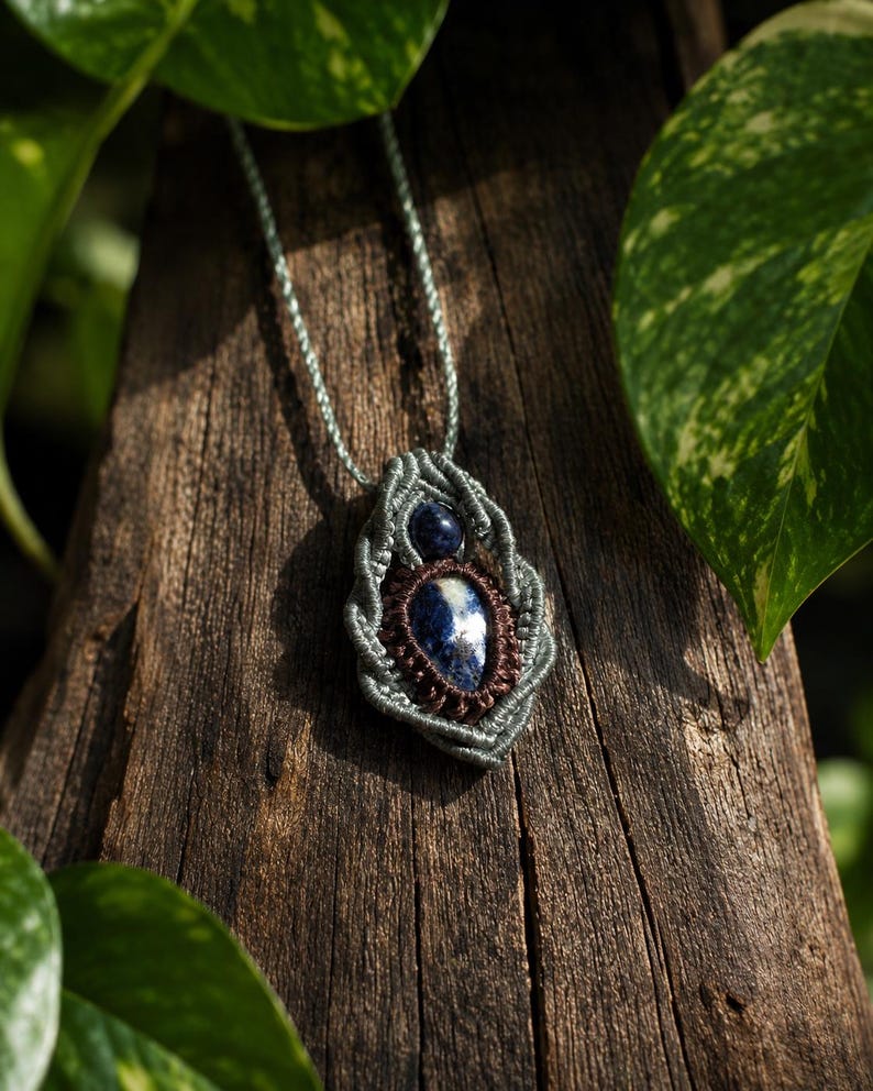 May include: A handmade macrame necklace featuring a blue and white stone pendant. The pendant is encased in a gray macrame design with a dark brown border. The necklace is displayed on a piece of weathered wood, surrounded by green foliage.