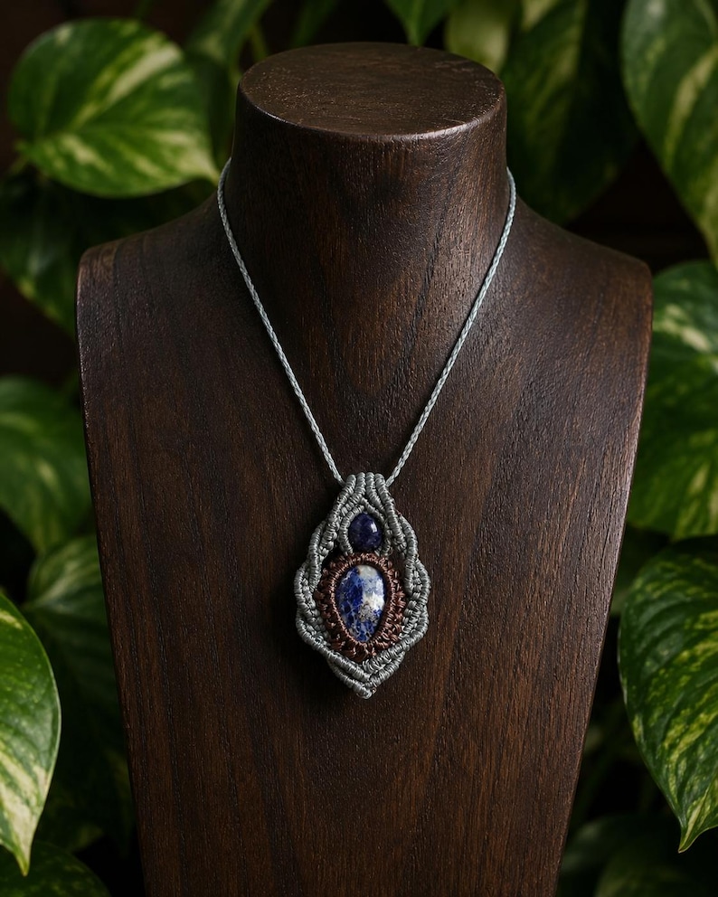 May include: A handcrafted macrame necklace featuring a large, oval-shaped blue gemstone pendant with a smaller blue stone above it. The pendant is encased in a gray macrame weave, suspended from a matching cord, displayed on a wooden bust.