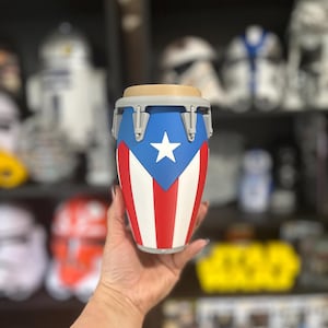 May include: A conga drum-shaped tumbler featuring the Puerto Rican flag design with a blue triangle, a white star, and alternating red and white vertical stripes. The top rim is tan, and the hardware accents are light gray.