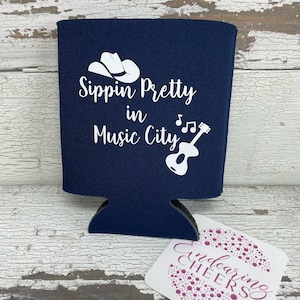 May include: A navy blue can cooler with white text that reads "Sippin' Pretty in Music City" with a cowboy hat and guitar graphic.
