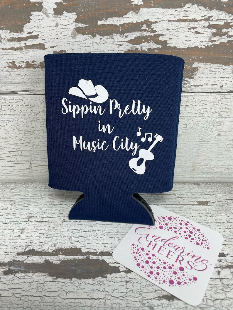 May include: A navy blue can cooler with white text that reads "Sippin' Pretty in Music City" with a cowboy hat and guitar graphic.