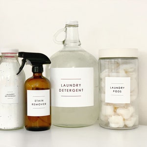 Puede incluir: Cuatro botellas de vidrio con etiquetas para detergente de ropa, quitamanchas, detergente en c&aacute;psulas y detergente de ropa. Las botellas est&aacute;n sobre una superficie blanca.