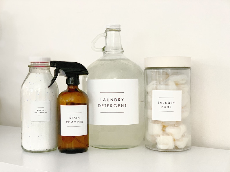 Puede incluir: Cuatro botellas de vidrio con etiquetas para detergente de ropa, quitamanchas, detergente en c&aacute;psulas y detergente de ropa. Las botellas est&aacute;n sobre una superficie blanca.