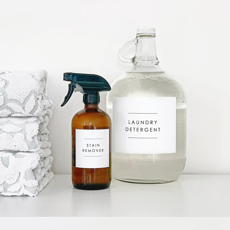 Puede incluir: Dos botellas de vidrio con etiquetas en un estante blanco. La botella de la izquierda es marr&oacute;n y tiene una boquilla de pulverizaci&oacute;n. La etiqueta dice "STAIN REMOVER". La botella de la derecha es transparente y tiene una etiqueta que dice "LAUNDRY DETERGENT".