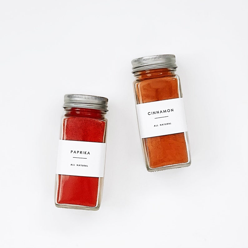 May include: Two glass jars with metal lids containing spices. The jar on the left contains paprika and the jar on the right contains cinnamon. Both jars have white labels with black text that reads "All Natural".