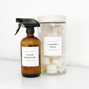 Puede incluir: Una botella de vidrio marr&oacute;n con una boquilla de pulverizaci&oacute;n negra etiquetada como "STAIN REMOVER" junto a un frasco de vidrio transparente con una tapa blanca lleno de pastillas de lavado blancas etiquetadas como "LAUNDRY PODS".