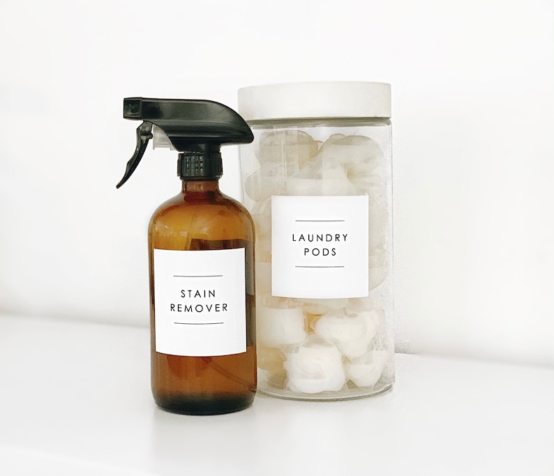 Puede incluir: Una botella de vidrio marr&oacute;n con una boquilla de pulverizaci&oacute;n negra etiquetada como "STAIN REMOVER" junto a un frasco de vidrio transparente con una tapa blanca lleno de pastillas de lavado blancas etiquetadas como "LAUNDRY PODS".