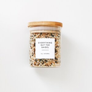 May include: A glass jar with a bamboo lid filled with a blend of sesame seeds, poppy seeds, dried onion, and garlic. The jar has a white label that reads "Everything But The Bagel" and "All Natural".